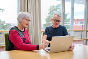 Punertavapaitainen nainen ja sinipaitainen mies tutkivat yhdessä tietokoneen käyttöä.